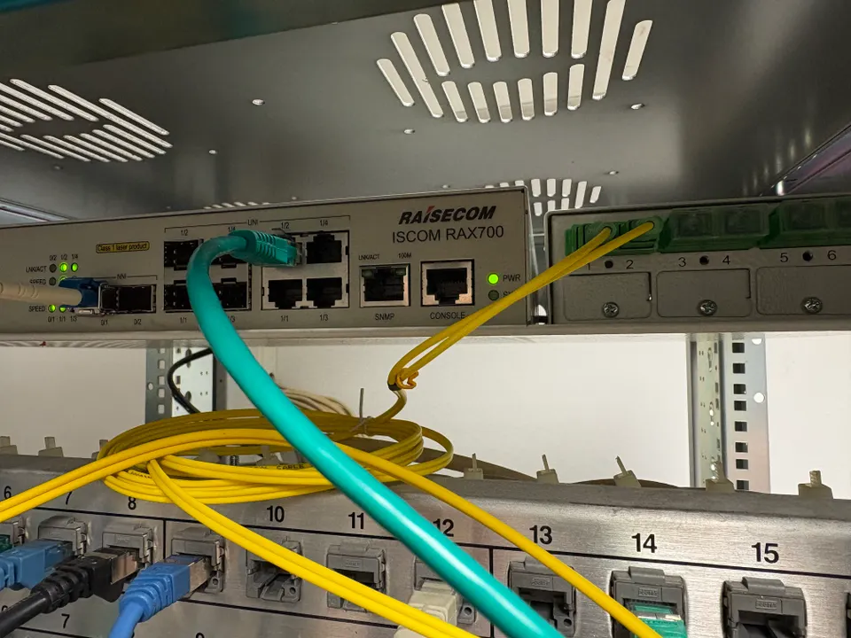 Une photographie présentant un équipement réseau de livraison client (CPE) de marque Raisecom (modèle ISCOM RAX700). Le port NNI 0/1 est connecté à une bi-fibre et le port UNI 1/2 est connecté à un câble réseau RJ45 vert. Les voyants d'état LNK/ACT 1/2 et LNK/ACT 0/1 sont allumés en vert fixe. Sous l'équipement, on peut voir le panneau de brassage de la baie.
